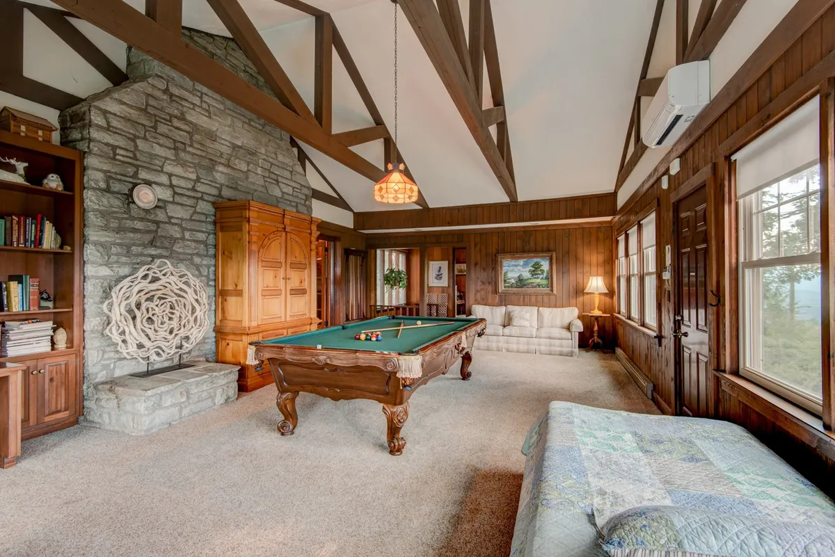 A spacious, warm gameroom featuring a pool table at its center, a stone fireplace to one side, built-in wooden bookshelves, large windows with scenic views, and comfortable seating areas. The mood is lively yet cozy, perfect for recreation and relaxation.