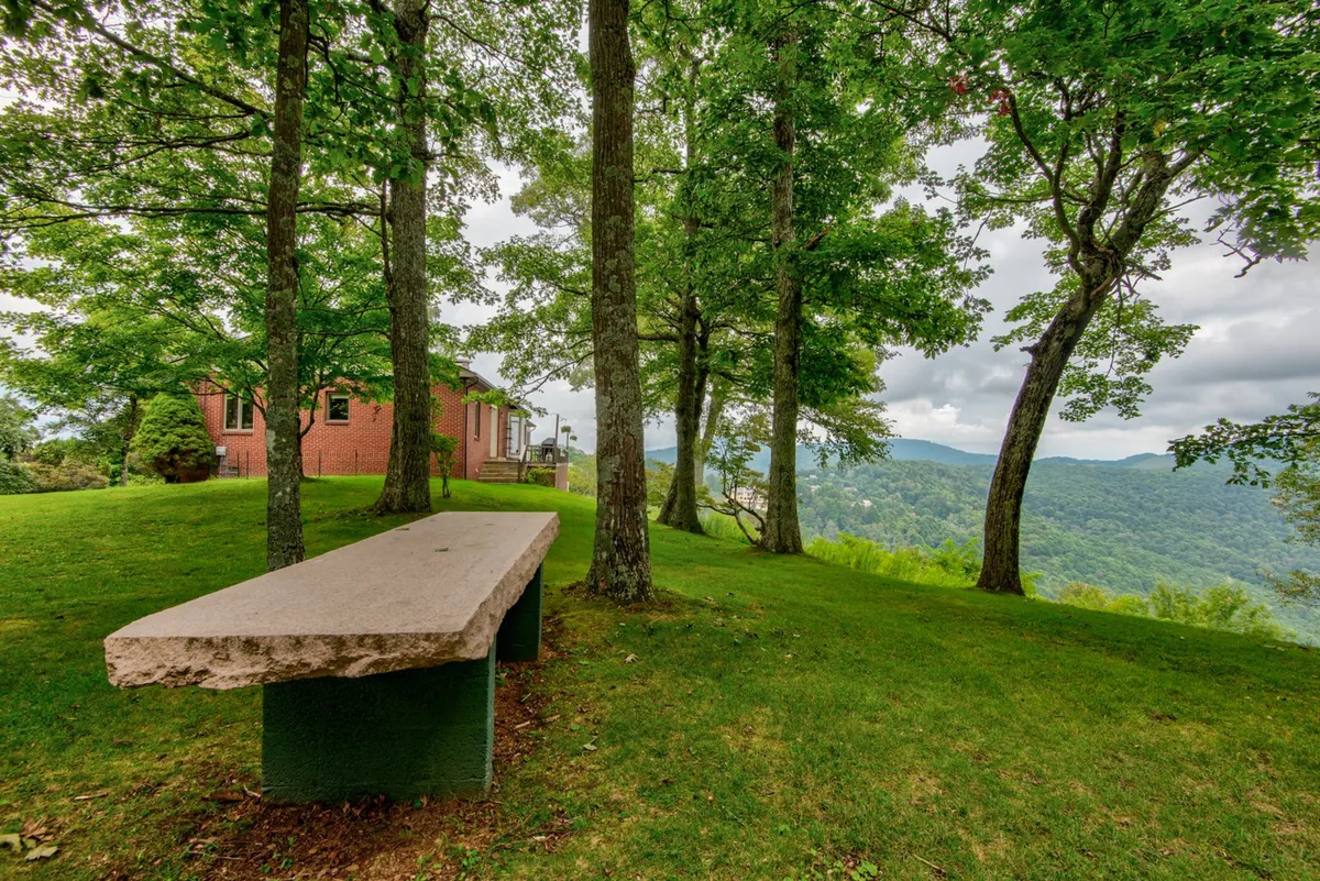 A tranquil stone bench sits amidst lush green grass under a leafy tree canopy, with distant mountain ranges completing the backdrop. This scenic spot evokes a peaceful mood and offers a versatile space for family relaxation, reading, or group gatherings.