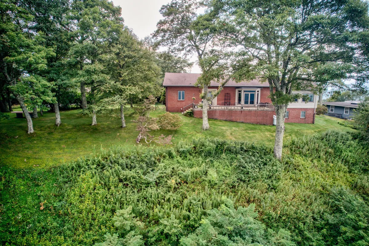 A peaceful exterior scene featuring a brick house with surrounding trees, a generous lawn, and a spacious deck area. The setting is ideal for group gatherings, evening sunset toasts, and quiet relaxation in nature.