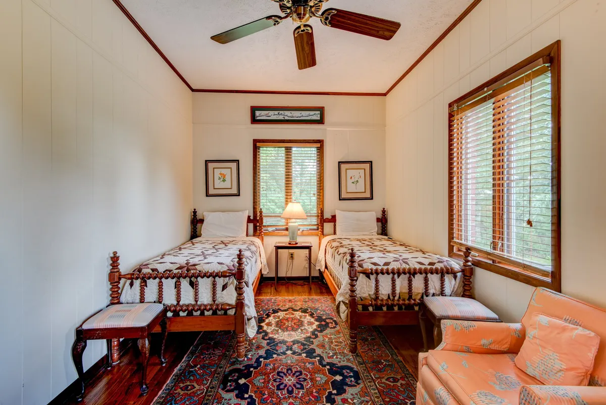 A welcoming bedroom with two twin beds neatly made, a soft area rug, an armchair, and a lamp between the beds. Natural light streams in through a nearby window, making the space feel inviting and ideal for siblings or friends.