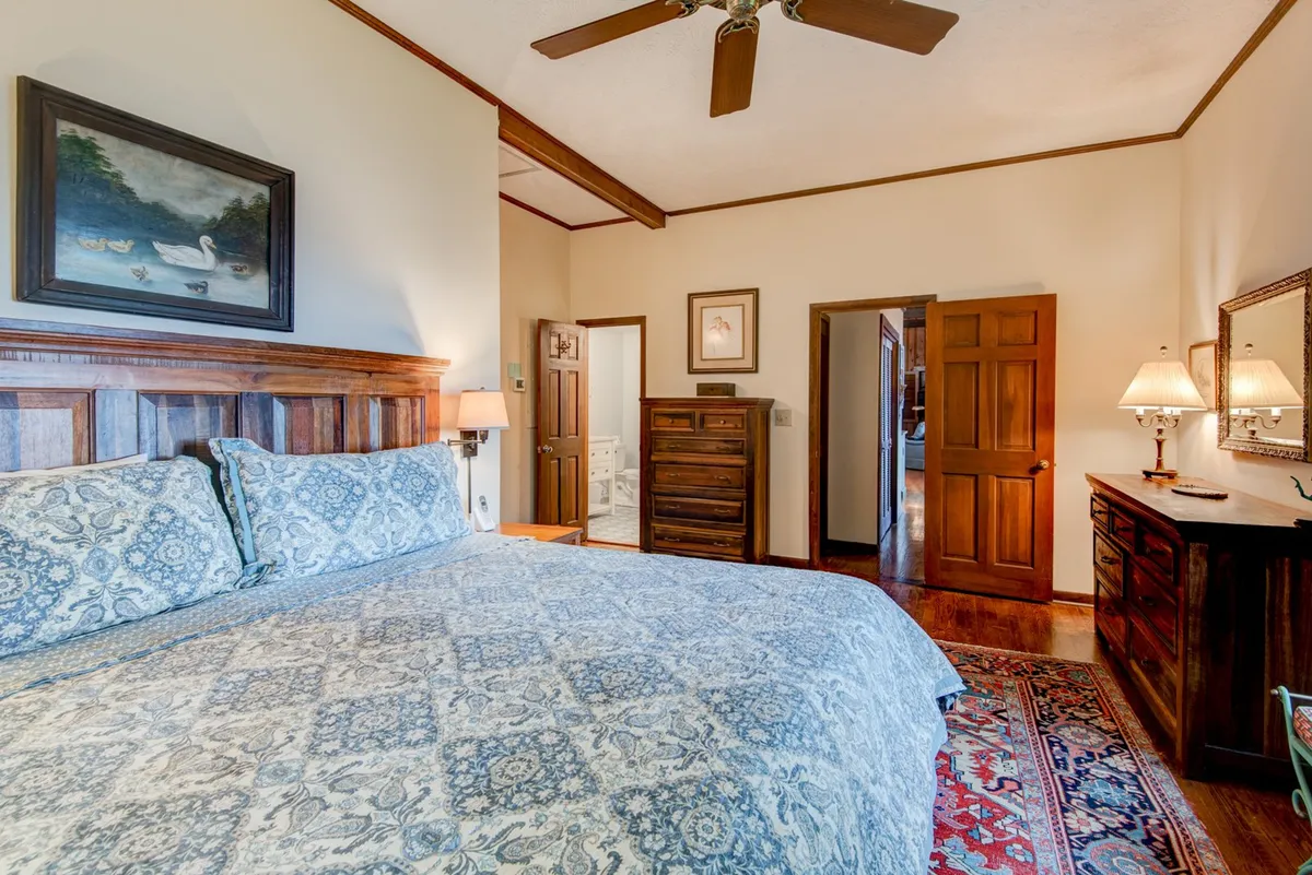 A serene master bedroom with a king-sized bed adorned in patterned bedding, flanked by wooden bedside tables and lamps, with elegant artwork above the headboard and intricate rugs underfoot. The space feels cozy, tranquil, and is bathed in gentle light, perfect for relaxation.