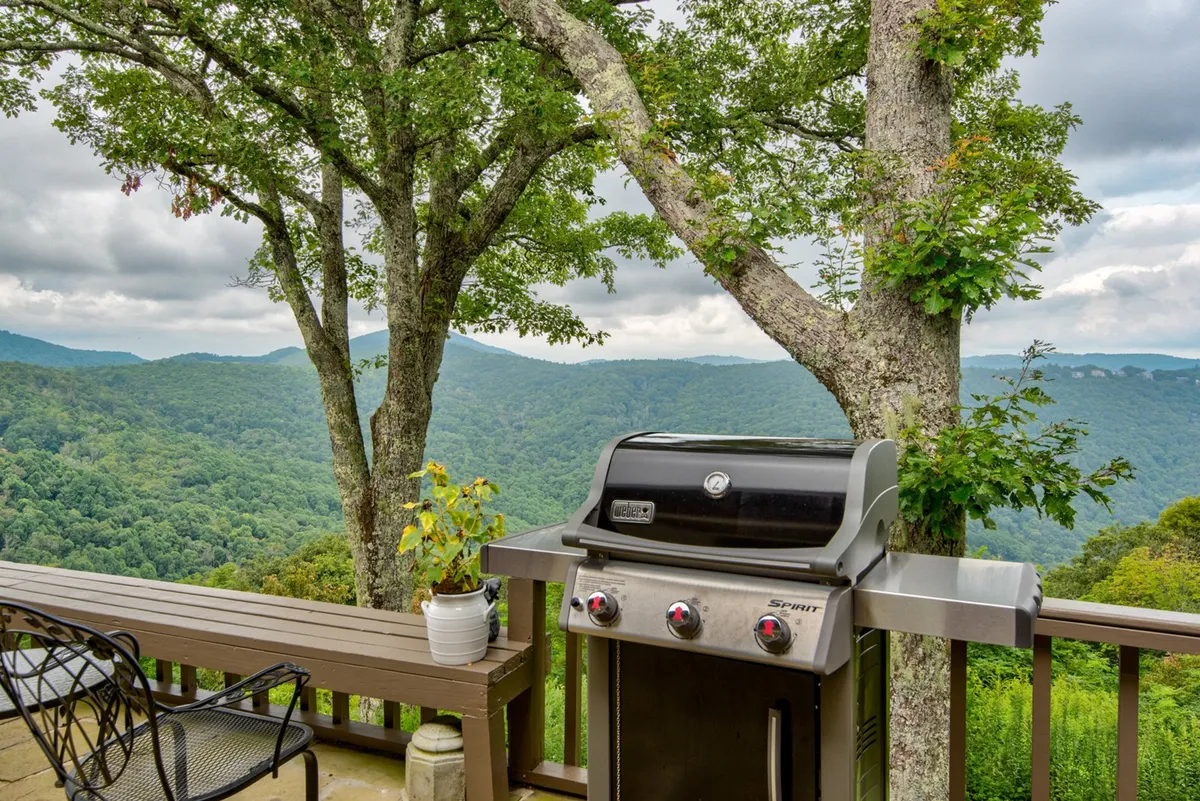 This exterior photo showcases a gas grill with patio furniture overlooking a sweeping mountain view framed by trees. The setting invites guests to enjoy grilling, outdoor dining, and relaxation, all while taking in the impressive scenery.