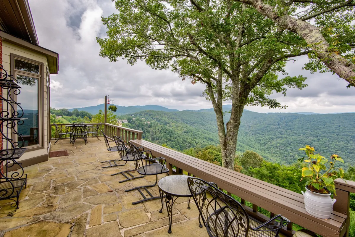 A peaceful patio with comfortable seating overlooks a stunning mountain vista. Stone flooring and a large tree add to the rustic mountain charm. The area is perfect for relaxing with a book, enjoying morning coffee, or hosting an outdoor meal while taking in panoramic views.