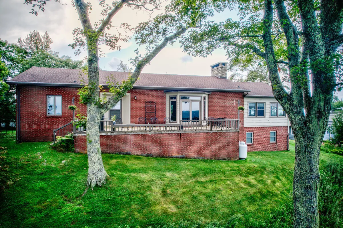 This image features the home's red brick exterior with a spacious deck surrounded by large windows and mature trees. The deck is set up for gatherings and relaxation, offering a clear view of the peaceful setting and the opportunity to host barbecues or watch sunsets.