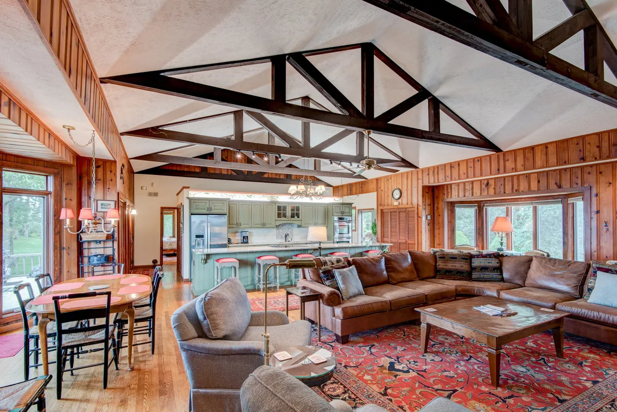 A spacious, open-plan living and dining area featuring a large sectional sofa, wooden dining table, kitchen island, exposed wooden beams, and expansive windows that showcase sweeping mountain views. The room exudes warmth and invites gatherings with its plush seating and seamless flow between lounging and dining spaces.