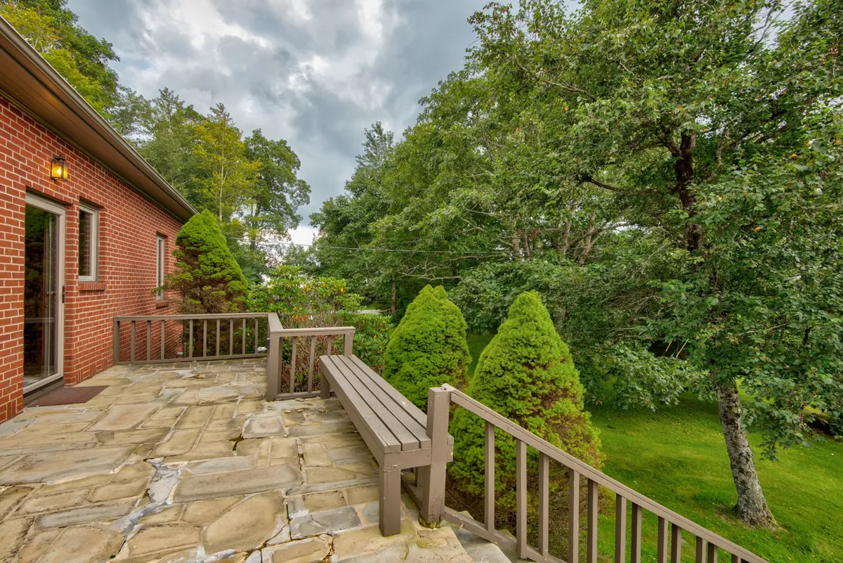 An inviting stone patio bordered by a wooden railing and lush green shrubs, set against a tranquil brick wall. The patio is ideal for relaxing, reading a book, or enjoying the fresh mountain air. The area feels open and welcoming, perfect for supervised play with pets or unwinding after a day outdoors.