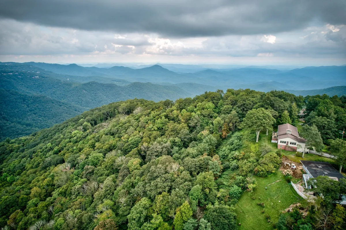 A peaceful exterior perspective of the house nestled among dense forest, with a panoramic view of the surrounding mountain range. The setting highlights the home's secluded mountaintop location, offering abundant space for outdoor activities like hiking or nature walks with pets.